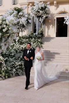 a bride and groom walking in front of a building with white flowers on the steps