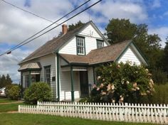 a white house with green trim and flowers on the front yard, next to a white picket fence