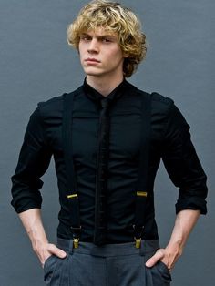 a young man with blonde hair wearing a black shirt and grey pants, standing in front of a gray background