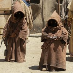 three men in brown robes standing next to each other on dirt ground with buildings in the background