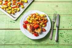 a plate with chicken and vegetables on it next to two trays of potatoes, carrots and peppers