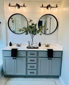 a bathroom with two sinks, mirrors and lights on the wall above them is a plant in a vase