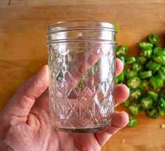 a person holding a glass jar filled with green peppers on top of a wooden table