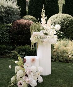 a tall white vase filled with flowers on top of a lush green field next to shrubbery