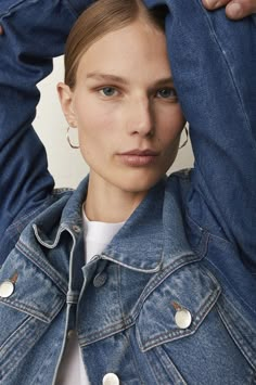 a woman in a denim jacket is holding her hands behind her head and looking at the camera