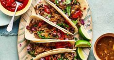 several tacos on a plate with sauce and spoons next to it, along with two bowls of salsa