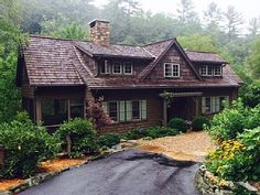 a house in the woods with lots of trees and bushes around it, along with gravel driveway