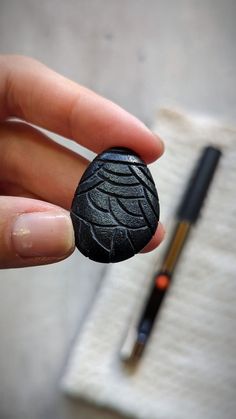 a hand holding a black rock with a pen in the background and a small piece of paper behind it