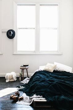 an unmade bed in a white room with two windows and a black blanket on the floor