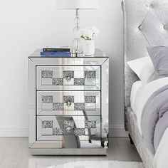 a mirrored chest of drawers with flowers on top in a white room next to a bed