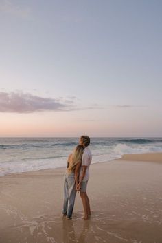 a man and woman standing on top of a sandy beach next to the ocean at sunset