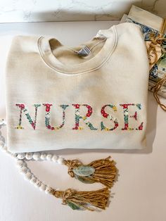 This embroidered floral font is absolutely beautiful. The detail to the flowers and colors are stunning.  Nurse embroidered to the front. Approximately 11x2 inches. Spring Cream Cotton Sweatshirt, Cream Cotton Sweatshirt With Letter Embroidery, Cream Sweatshirt With Embroidered Text For Fall, Embroidered Long Sleeve Sweatshirt For Spring, Spring Sweatshirt With Letter Embroidery And Long Sleeves, Fall Cotton Sweatshirt With Floral Embroidery, Spring Long Sleeve Sweatshirt With Letter Embroidery, Spring Floral Embroidery Long Sleeve Sweatshirt, Long Sleeve Sweatshirt With Letter Embroidery For Spring