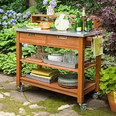 an outdoor kitchen cart with wine glasses, plates and bottles on it in front of some plants