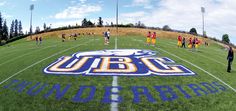 a group of people standing on top of a field next to a football field with the ubc logo painted on it