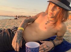 a shirtless man sitting on the beach with a cup in his hand and wearing a fedora