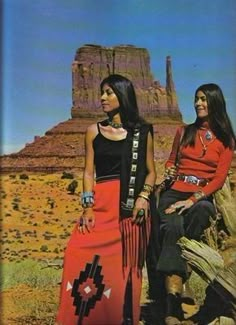 two women in native american clothing standing next to each other on a desert landscape with rocks and mountains in the background