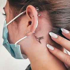 a woman has a small bird tattoo on her left side behind the ear and behind the ear is a pair of hoop earrings