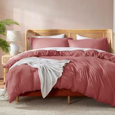 a bed with pink sheets and pillows in a bedroom next to a potted plant