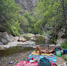 a woman laying on top of a blanket next to a river in the woods with a camera