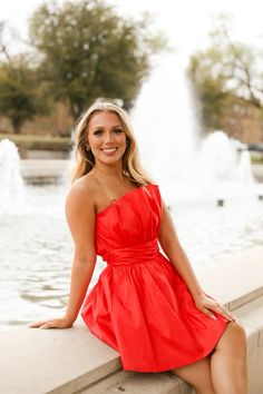 a woman in a red dress is sitting on a ledge near a fountain and smiling at the camera