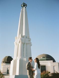 a man and woman standing in front of a tall white monument with a clock on top