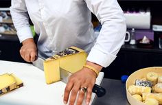 a man in white shirt cutting cheese on top of a counter next to other food items