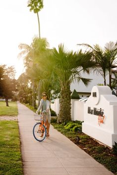 a woman riding a bike down a sidewalk next to palm trees and a white sign