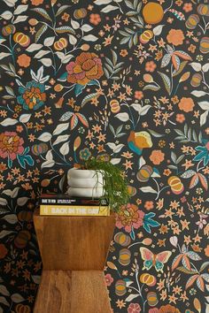 a wooden table topped with books next to a wall covered in floral wallpaper and a potted plant