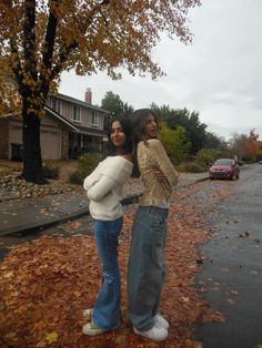 two women standing on the side of a road in front of trees with leaves all over them