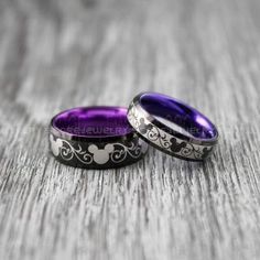 two wedding bands with mickey mouse designs on them sitting on a wooden surface, one is purple and the other is black