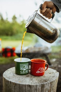 a person pouring coffee into two mugs on top of a tree stump in front of a campfire