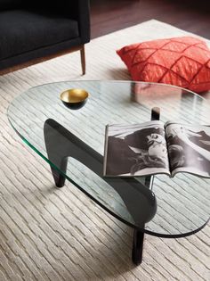 a glass coffee table with an open magazine on it's top and a red pillow in the background