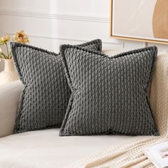 two gray pillows sitting on top of a white couch next to a vase with flowers