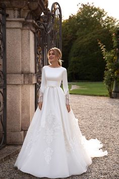 a woman in a white wedding dress standing by an iron gate with her hands on her hips