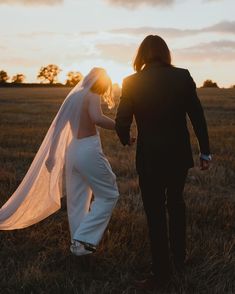 a bride and groom holding hands walking through a field at sunset with the sun behind them