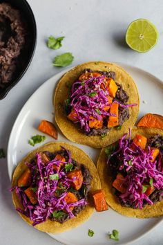 three tacos on a white plate with limes and cilantro in the background