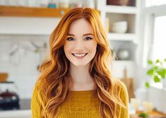 a woman with long red hair standing in a kitchen smiling at the camera while wearing a yellow sweater