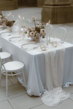 the table is set with white chairs and flowers on it, along with candles in vases