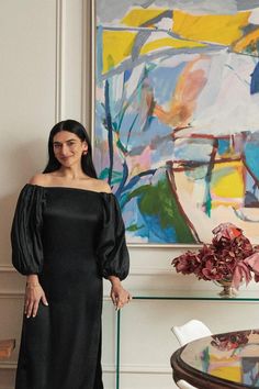 a woman standing next to a painting in a living room with a glass table and chairs