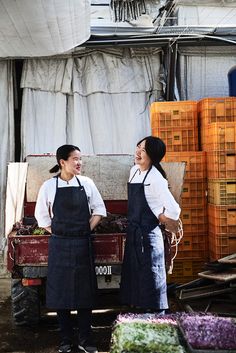 two women in aprons standing next to each other near crates and boxes on the ground