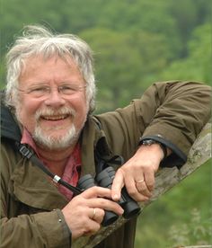 an older man with grey hair and beard holding a camera in his hands while leaning on a tree branch