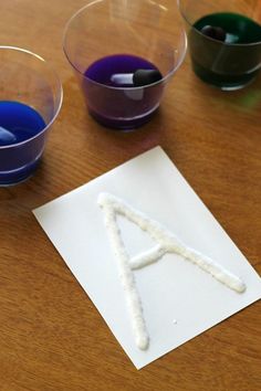 three different colored liquids in small bowls on a table with one letter made out of sticks