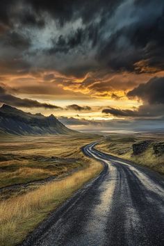 an empty road in the middle of nowhere with mountains in the background and clouds overhead