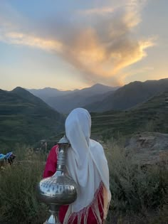 a woman in traditional garb carrying a silver vase on top of a hill with mountains in the background