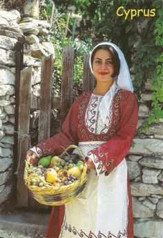 a woman in traditional dress holding a basket of fruit next to a stone wall and fence