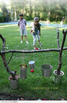 two children playing with tin cans on a tree branch in the grass, and one child is holding an apple