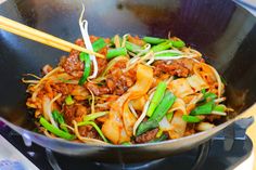 a wok filled with noodles and vegetables on top of a stove burner next to chopsticks