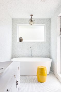 a white bath tub sitting under a window next to a yellow foot stool in a bathroom