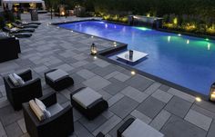 an outdoor swimming pool surrounded by lounge chairs and lights at night with water feature in the background