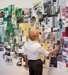 a woman standing in front of a wall covered with pictures and papers, pointing at something on the wall